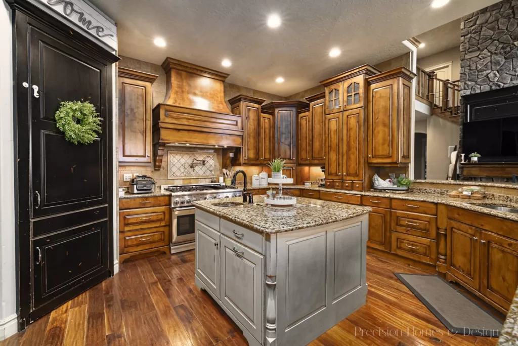 PHD interior kitchen design. Wood floor, spacious granite counters with cooking island, black pantry door, and plenty of cabinet space in brown wood finish.
