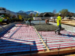 Construction site pouring concrete foundation with radiant hydronic heating system and workers in safety gear.