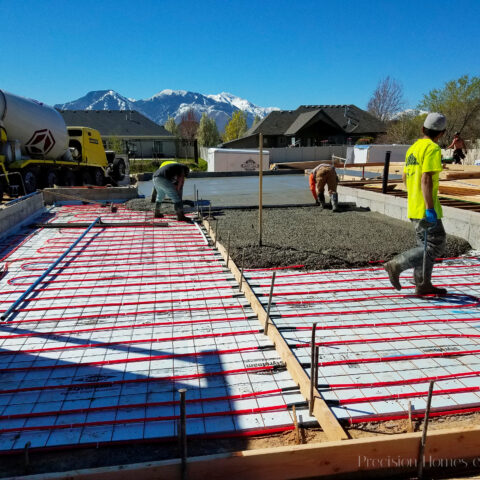 Construction site pouring concrete foundation with radiant hydronic heating system and workers in safety gear.